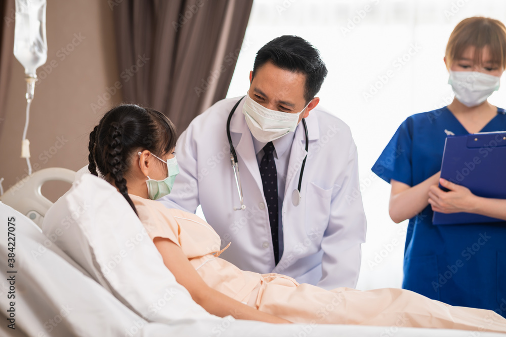 Asian male doctor consoling and talking with recovering girl patient during medical visit at hospital ward with female nurse standing in background. Medical workers visiting a girl patient. Asian male doctor consoling and talking with recovering girl patient during medical visit at hospital ward with female nurse standing in background. Medical workers visiting a girl patient.