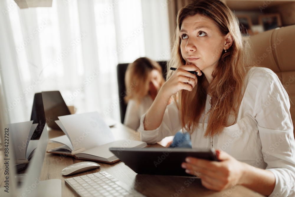 Concentrated woman sit at the desk, hold tablet, thinking over, making decisions, young mother working at home online, while caring grandmother babysit with newborn baby girl Concentrated woman sit at the desk, hold tablet, thinking over, making decisions, young mother working at home online, while caring grandmother babysit with newborn baby girl