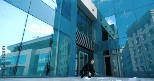 Young clumsy office manager lifting the fallen documents from floor near modern business center. Trainee manager late for work for interview or conference meeting, loses papers on way, feels despair