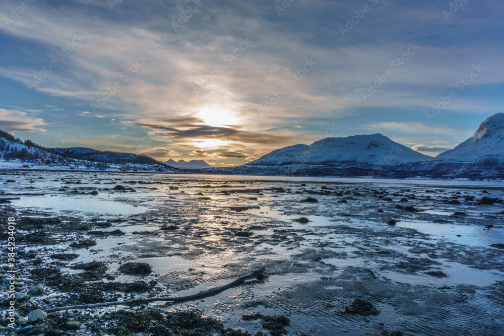 Beach at low tide in Norway with snowcaped mountains in the distance, sunlight reflecting in the water 