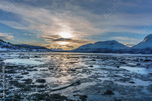 Beach at low tide in Norway with snowcaped mountains in the distance, sunlight reflecting in the water 