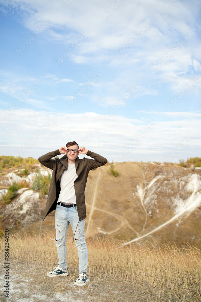 Man with glasses in jeans, t-shirt and cape stands in hilly terrain. Adult male in hood adjusts his eyeglasses and enjoys beautiful view of countryside in windy weather. Slow motion.