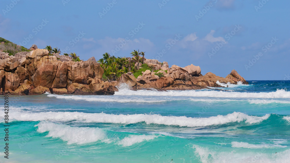Fototapeta premium Characteristic beautiful granite rock formations with coconut trees in between and breaking waves in turquoise water on beach Petite Anse in the south of La Digue island, Seychelles.