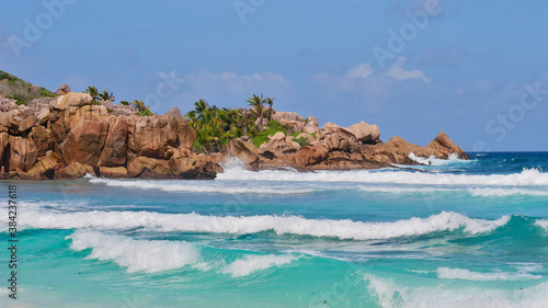 Characteristic beautiful granite rock formations with coconut trees in between and breaking waves in turquoise water on beach Petite Anse in the south of La Digue island, Seychelles.