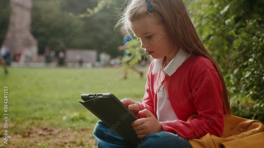 Adorable kid beautiful girl with blonde hair using tablet computer studying online doing school lessgons sitting on grass outdoors. Distant education. Elementary school. Adorable kid beautiful girl with blonde hair using tablet computer studying online doing school lessgons sitting on grass outdoors. Distant education. Elementary school.