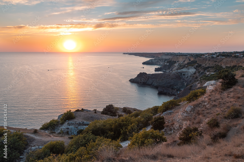 Panoramic landscape at the Cape Fiolent during the sunset. A large crimson Sun sets over the horizon against a cloudy sunset pink sky. Twilight sun shines on rocky cliffs. Black sea below the cliffs.