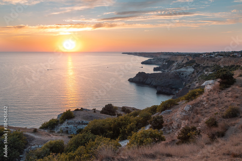 Panoramic landscape at the Cape Fiolent during the sunset. A large crimson Sun sets over the horizon against a cloudy sunset pink sky. Twilight sun shines on rocky cliffs. Black sea below the cliffs. © Vadim