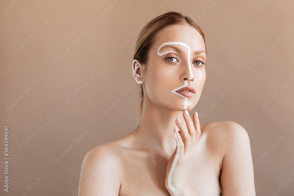 Young woman without makeup with clean skin looks into camera on beige background. Portrait of girl with white stripe on her face and hand Young woman without makeup with clean skin looks into camera on beige background. Portrait of girl with white stripe on her face and hand