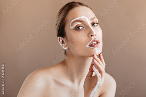 Young woman with white line in middle of her face touches her chin and sensitively looks into camera on beige background photo