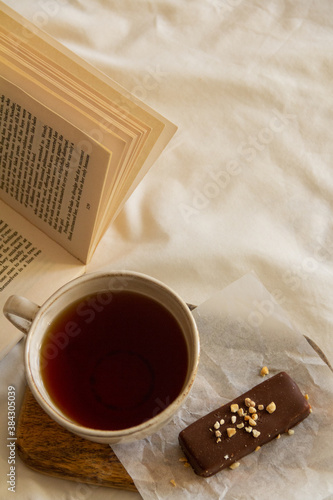 Cup with tea and a chocolate bar on a wooden board next to an open book on a white bed