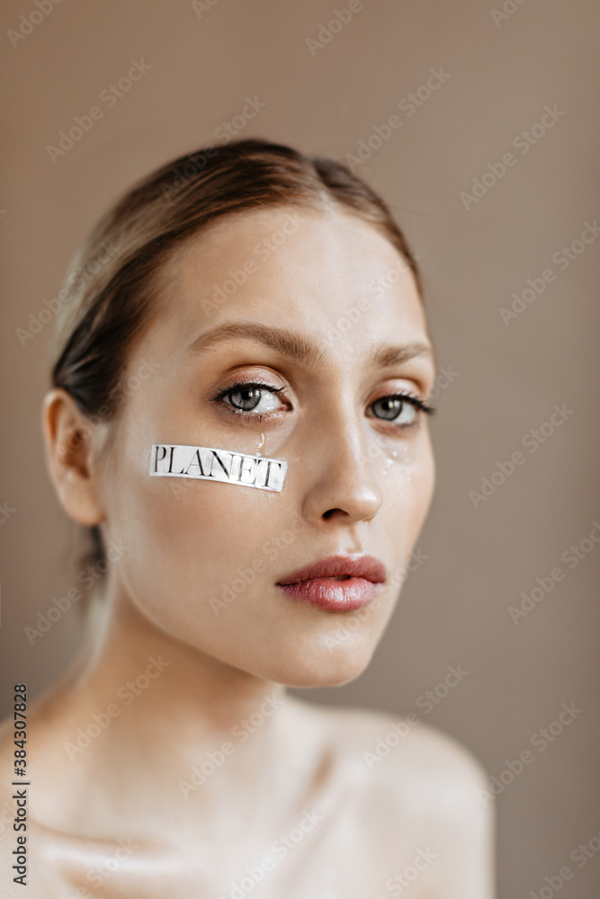 Naklejka premium Saddened woman with word planet on her cheek posing on beige background. Girl with gray eyes is crying