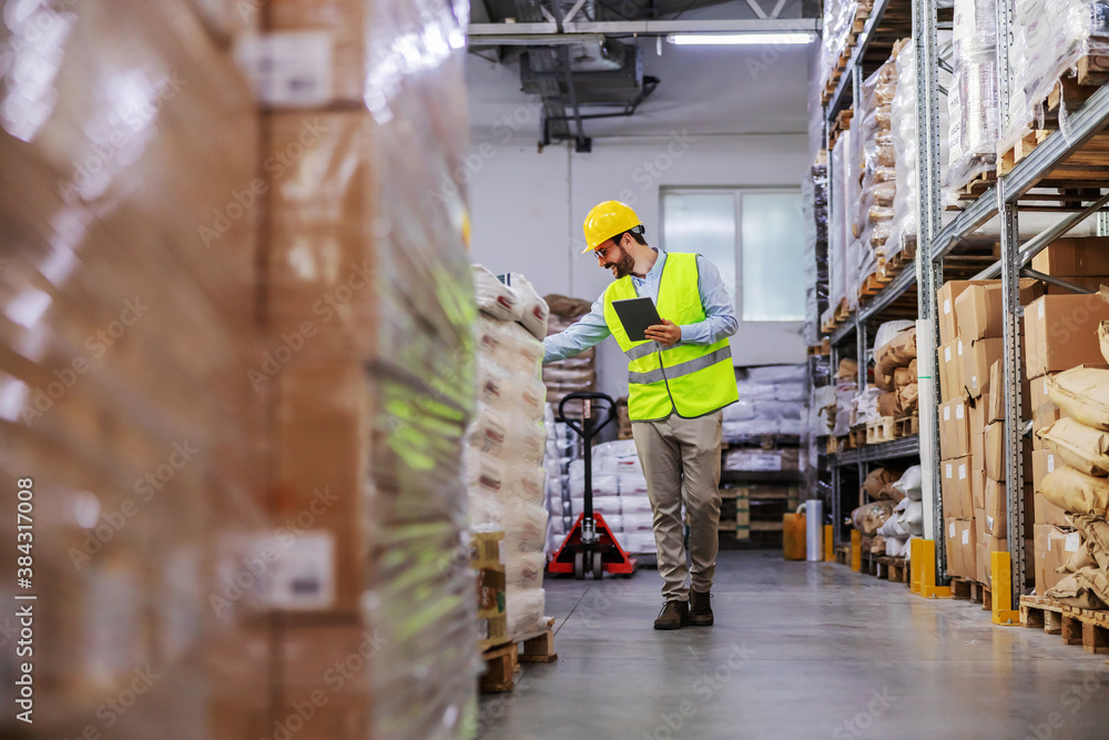Full length of attractive bearded supervisor walking around warehouse and using tablet to check on goods for shipment. - obrazy, fototapety, plakaty 