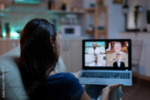 Woman in pijamas lying on sofa using laptop talking about sale report in video conference with team. Remote worker having online meeting consulting with colleagues using videocall and webcam chat