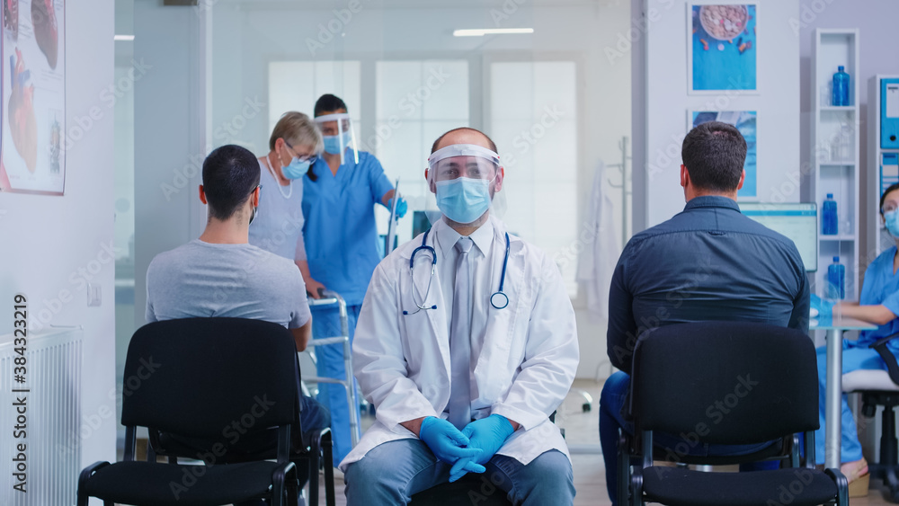 Doctor with visor against coronavirus in waiting area looking intro camera. Nurse with face mask assisting disabled woman with walking frame in hospital hallway. Doctor with visor against coronavirus in waiting area looking intro camera. Nurse with face mask assisting disabled woman with walking frame in hospital hallway.