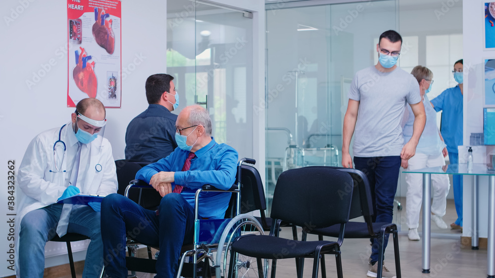 Invalid old man with face mask against infection with coronavirus in wheelchair discussing with doctor in hospital waiting area. Nursing inviting patient in examination room.