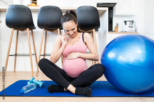 Young pregnant female working out in living room. Caucasian woman in a sports outfit practicing fitness at home. Using cell phone. Young mom has on conversation with family. Kitchen in the background.