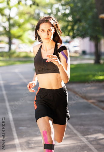 Front view of attractive young caucasian professional bodybuilder with elastic kinesiology tapes on body running at stadium. Woman with muscular perect body looking at camera. Workout concept. photo