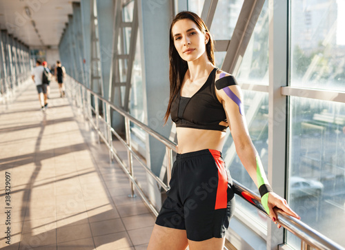 Portrait of muscular brunette woman wearing black sports outfit, looking at camera. Young smiling female athlete posing near handrails indoors, colorful kinesiotaping on body, futuristic interior.