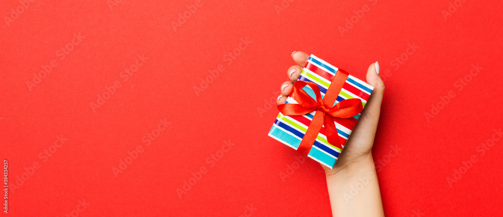 Female's hands holding striped gift box with colored ribbon on red background. Christmas concept or other holiday handmade present box, concept top view with copy space