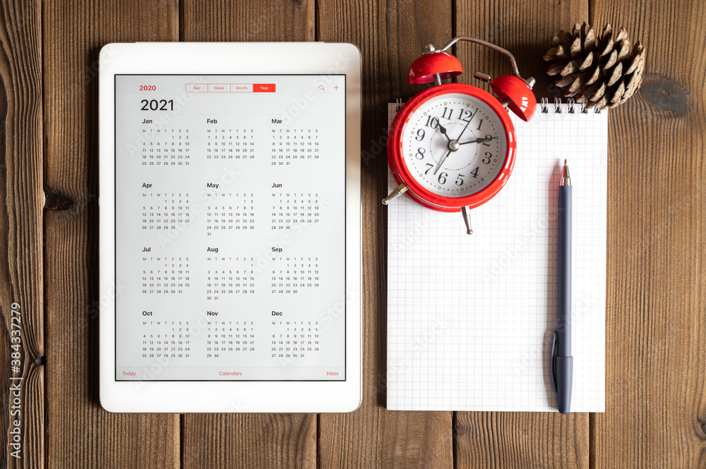 a tablet with an open calendar for 2021 year, a red alarm clock, a pine cone, and a spring notebook with a pen on a wooden boards table background a tablet with an open calendar for 2021 year, a red alarm clock, a pine cone, and a spring notebook with a pen on a wooden boards table background