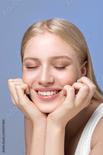 Studio shot of young blonde woman smiling with teeth isolated on a blue background. Happy beautiful girl. Closed eyes and broad smile. Skin care, beauty, wellness, health concept