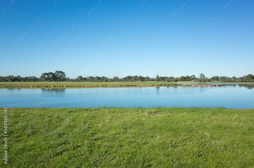 Grass, lake, and lookout platform in the distance against the cloudless blue sky. Background texture of the Australian nature landscape of a large public park. Presidents Park lake, VIC Melbourne.