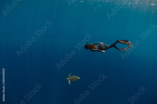 A female freediver is swimming with a sea turtle in a blue ocean.