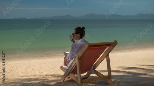  woman drinks coffee at the tropical beach resort, she seats by the sea line