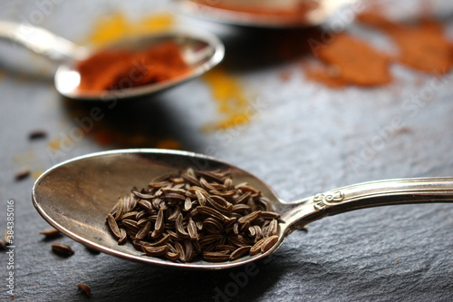 A close up photograph of caraway seeds on a silver teaspoon against a slate counter worktop.  photo