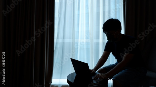 A young asian man with a mask in a silhoutte is working with his laptop in his home (work from home)