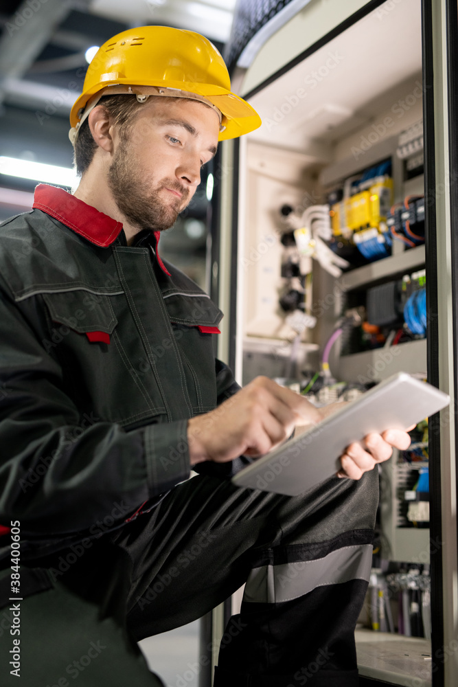 Young bearded engineer in workwear and hardhat checking settings of machine Young bearded engineer in workwear and hardhat checking settings of machine