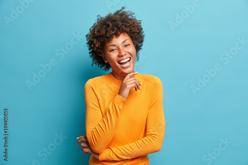 Portrait of glad young Afro American woman laughs happily keeps hand on chin expresses positive emotions smiles broadly has carefree expression wears orange jumper isolated over blue background