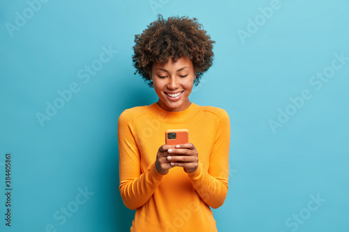 Online lifestyle concept. Cheerful good looking woman with Afro hair sends text messages via mobile phone dressed casually searches gifts for holiday in internet uses smartphone app browses webpage