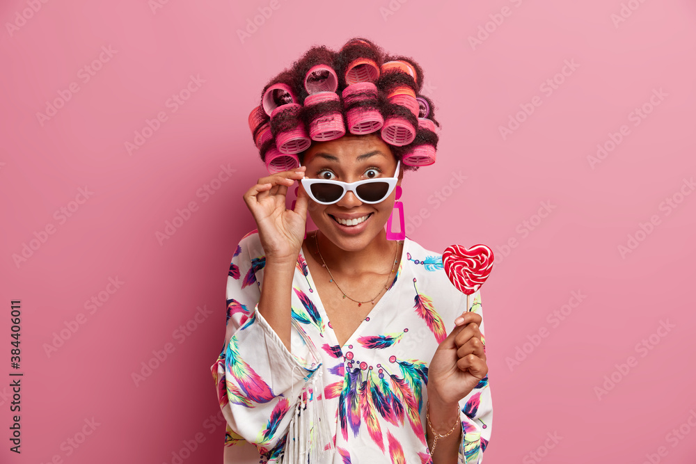 Positive dark skinned female model keeps hand on sunglasses wears hair rollers for making perfect hairdo enjoys domestic atmosphere holds candy isolated on pink background. Women style concept