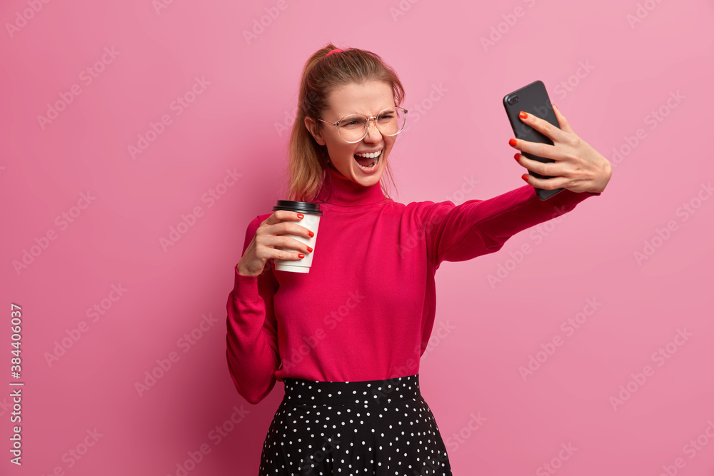 Angry emotional girl takes selfie on smartphone holds takeaway coffee poses with unhappy grimace dressed in stylish clothes against pink background. Displeased teenager makes photo of herself Angry emotional girl takes selfie on smartphone holds takeaway coffee poses with unhappy grimace dressed in stylish clothes against pink background. Displeased teenager makes photo of herself