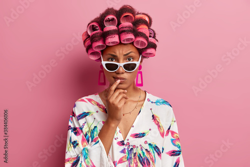 Serious strict dark skinned woman wears sunglasses and dressing gown makes curly hairstyle with hair rollers thinks about her image on party poses indoor against pink background. Beauty concept © wayhome.studio 