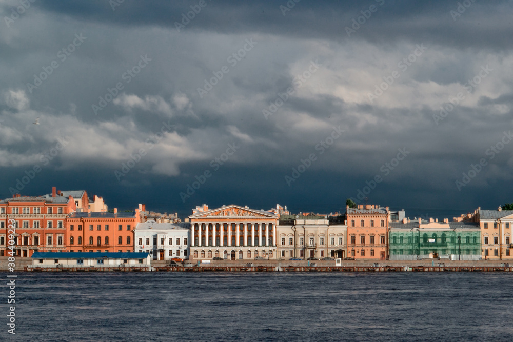 Embankment of Saint Petersburg city with colorful bright red white houses on banks of blue Neva River in the light of sunset. Dark storm clouds