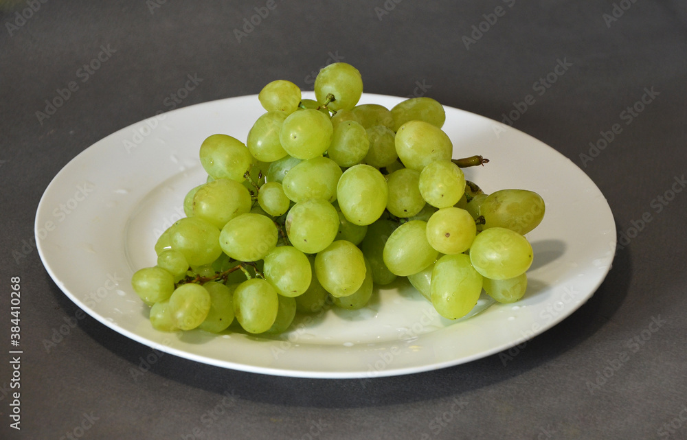 Juicy green grapes on a white plate. Delicious fresh fruits on a gray background. Juicy green grapes on a white plate. Delicious fresh fruits on a gray background.