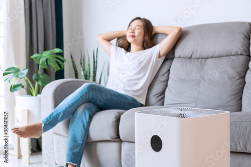 Pretty young Asian woman on comfortable sofa, hands behind head rest at home wellbeing breathing fresh air from air purifier while dust air pollution situation outside is r
