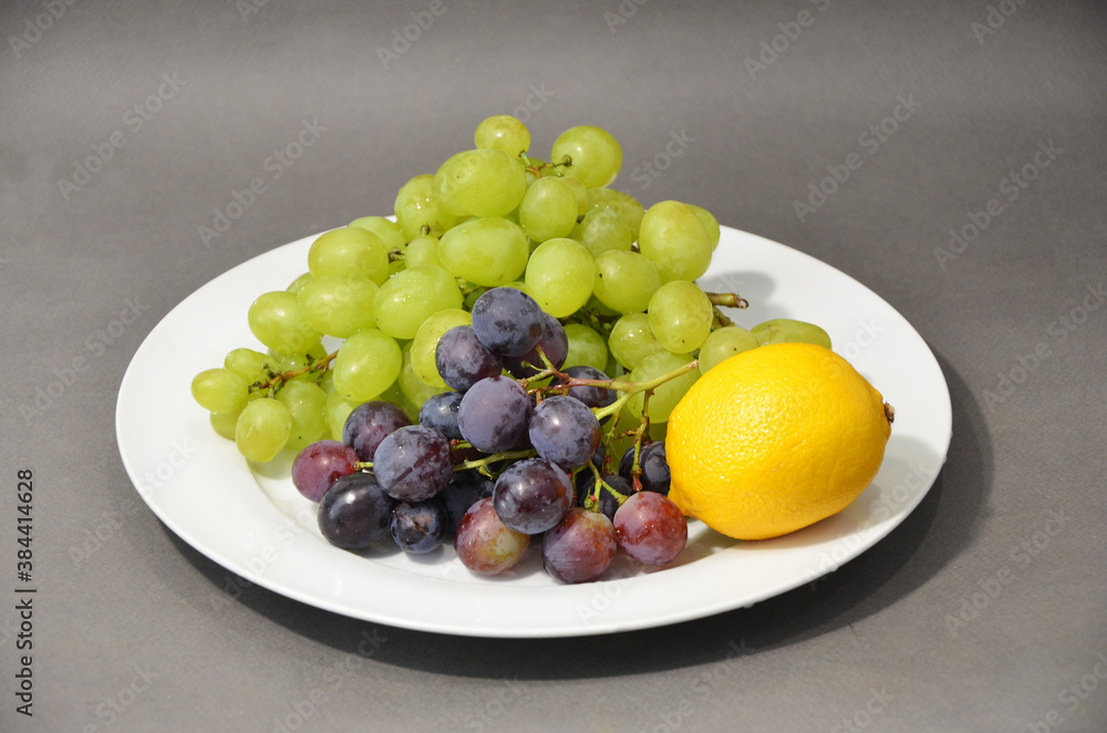Multi-colored grapes and yellow lemon laying on a white plate. Fruit on a gray background. Multi-colored grapes and yellow lemon laying on a white plate. Fruit on a gray background.