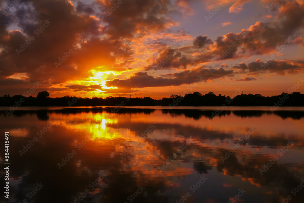 Amazing sunrise in rural scene. Symmetry of the sky in a lake at sunset. Clouds reflecting on the water. Quiet relaxing scene with a beautiful colorful cumulonimbus. Silhouette of vegetations.