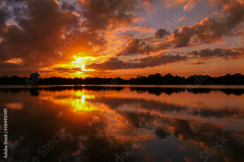 Amazing sunrise in rural scene. Symmetry of the sky in a lake at sunset. Clouds reflecting on the water. Quiet relaxing scene with a beautiful colorful cumulonimbus. Silhouette of vegetations.