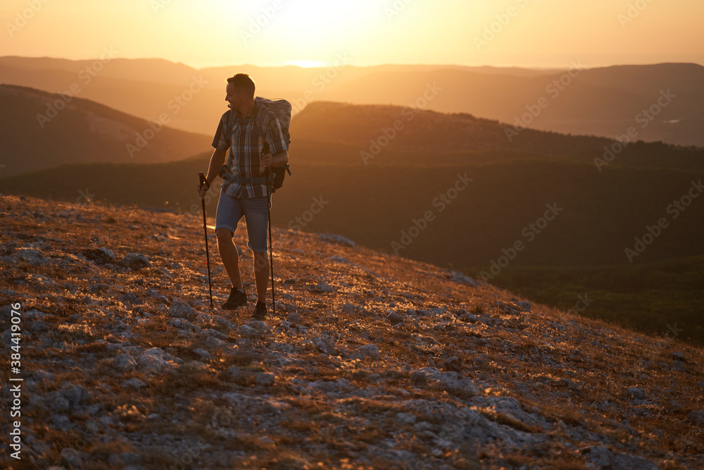Young hiker with backpack and trekking poles walking along a mountain path at sunset with a smile. High quality photo.