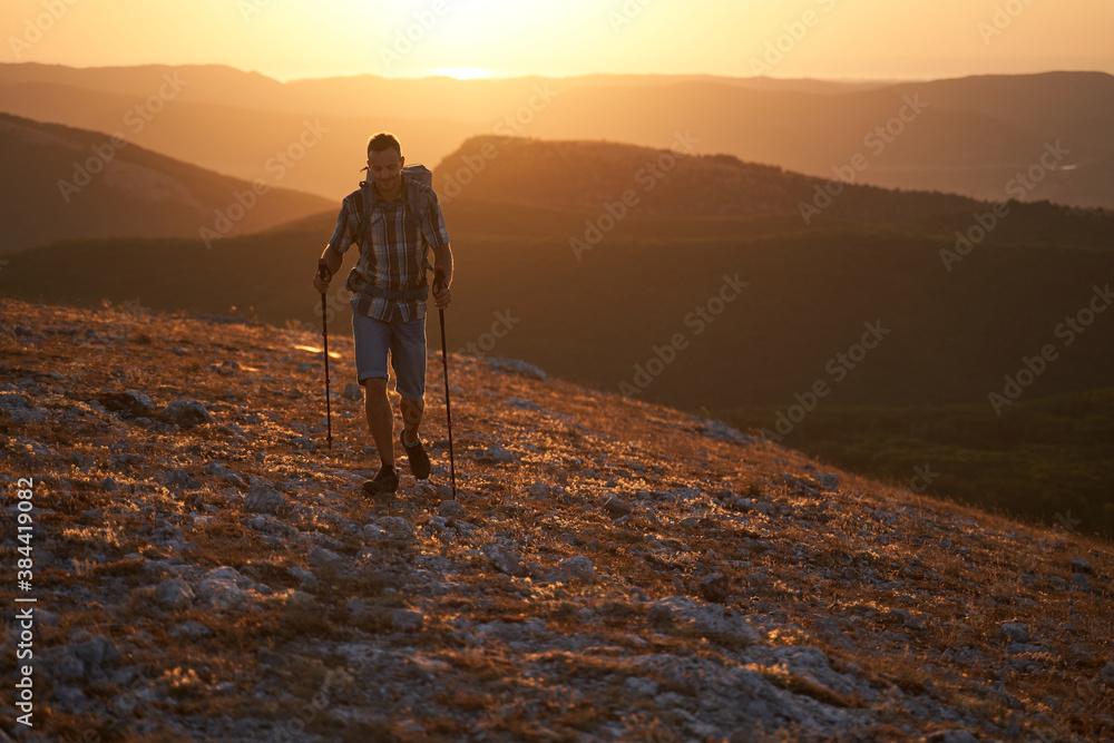 Young hiker with backpack and trekking poles walking along a mountain path at sunset with a smile. High quality photo.