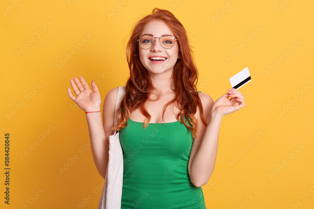 Young Caucasian woman with shopping bag and blank credit card posing with spreading hands and excited facial expression, wearing green t shirt, has red wavy hair. Young Caucasian woman with shopping bag and blank credit card posing with spreading hands and excited facial expression, wearing green t shirt, has red wavy hair.