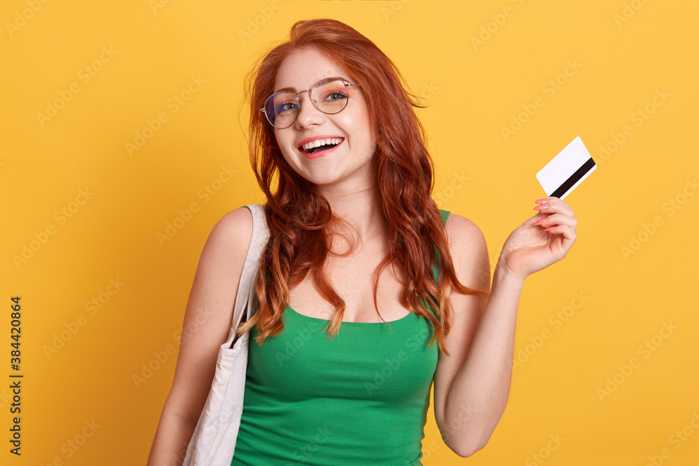 Naklejka premium Portrait of woman holding shopping bag and credit card isolated over yellow background, red haired female being ready to do shopping, excited lady enjoying her purchases.