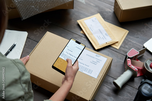 Female warehouse worker, seller, dropshipping small business owner holding phone scanning retail package postal parcel barcode on ecommerce shipping box label on smartphone using mobile app, close up. photo