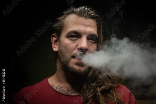 Close up of a young man exhaling a cloud of steam or smoke with an electronic cigarette.