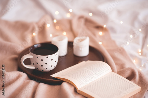 Cup of tea with open book and candles on wooden tray close up over Christmas lights in bed. Winter holiday season. Good morning. Breakfast.