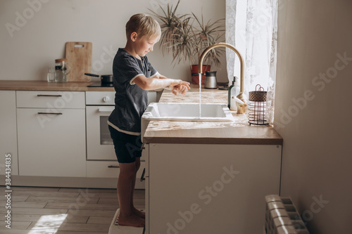A boy of European appearance washes his hands in a light kitchen at home. 